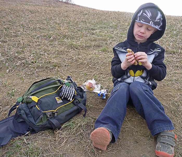 Kid snacking on the river bank in Denver, while on a fly fishing trip with his dad, sitting next to a Mountainsmith Tour TLS Lumbar Pack.