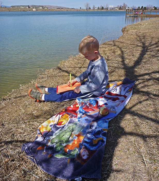 Child site on the river bank in Denver coloring and snacking while on a fly fishing outing with his dad.