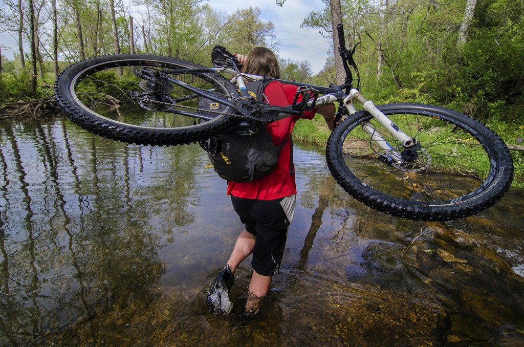 Curtis Savard wades through water with his mountian bike over his shoulder and a Mountainsmith Wraith 25 backpack on, image by aaron codling