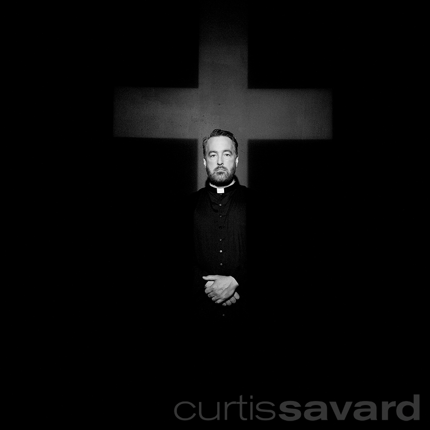 black and white photography by curtis savard of a priest in front of a cross
